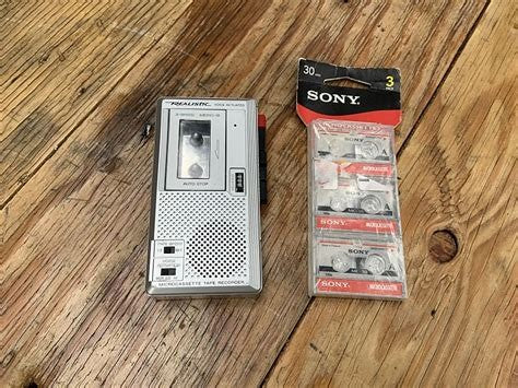 A Realistic microcassette recorder next to a sealed 3-pack of Sony microcassettes
